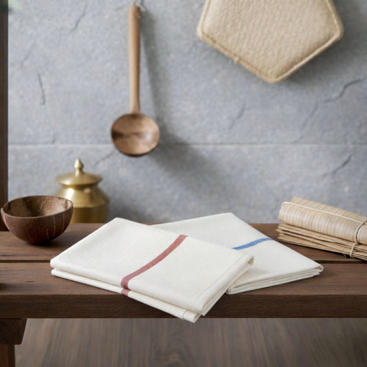 zv-pack=set-of-2 | Two folded ZARUVA Kerala thorthu mundu bath towels on a wooden surface in a traditional Indian bathroom setting — white flat-weave (non-terry) 100% cotton with blue and maroon borders 